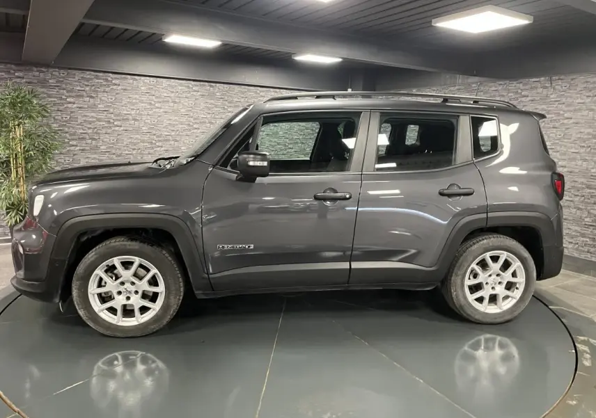 Vue de profil côté gauche d'un Jeep Renegade gris métallisé avec barres de toit noires en intérieur showroom.