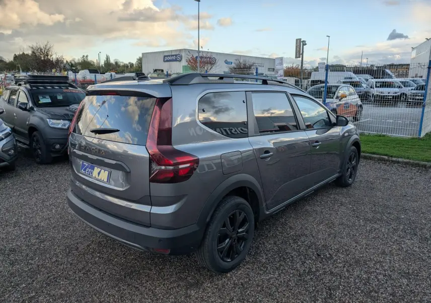 Vue 3/4 arrière droite d’un Dacia Jogger gris métallisé avec jantes noires et barres de toit, stationné sur un sol gravillonné.