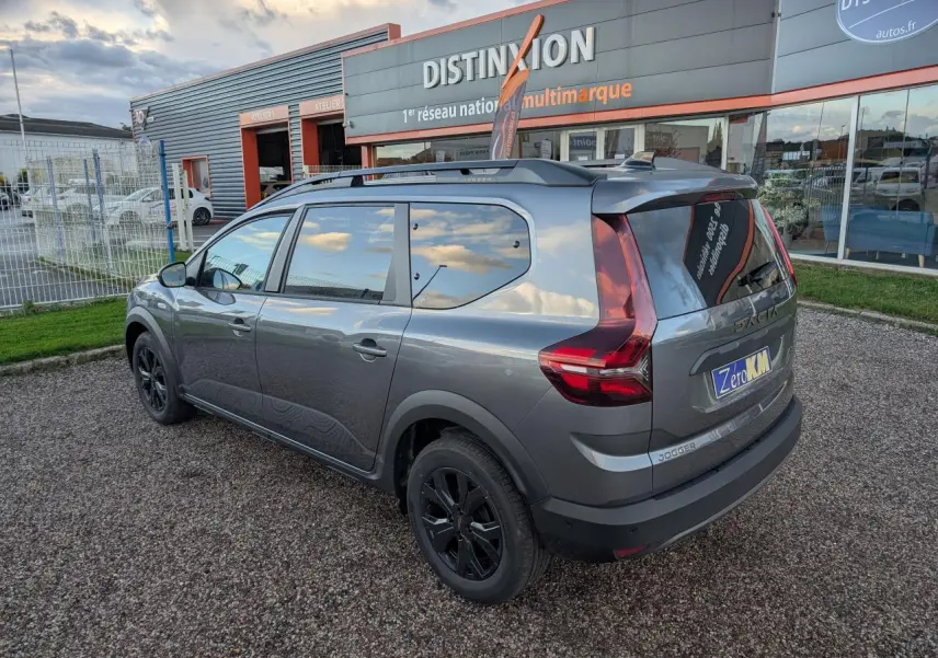 Vue 3/4 arrière droite d'un Dacia Jogger gris métallisé avec jantes noires et barres de toit, stationné devant un concessionnaire.