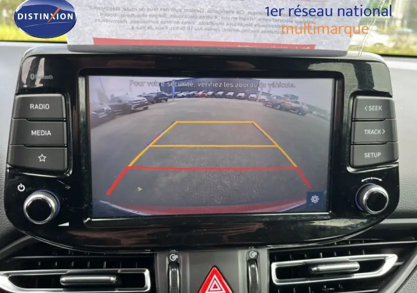 Vue intérieure de l’écran multimédia affichant la caméra de recul d’une Hyundai i30 SW rouge, avec commandes autour de l’écran.