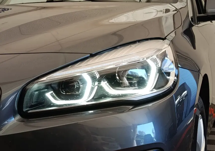 Gros plan sur le phare avant droit allumé d'une BMW Série 2 Gran Tourer F46 gris clair, reflet sur la carrosserie.