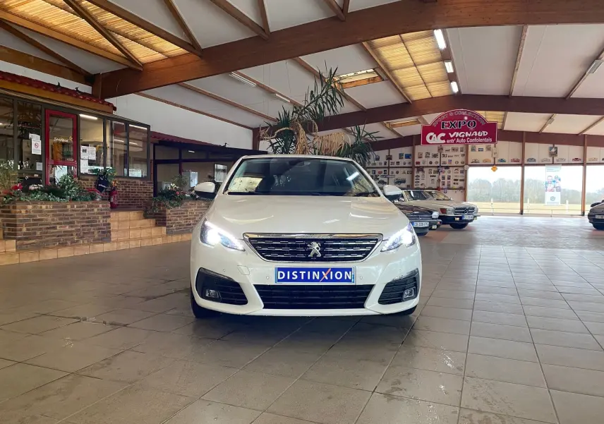 Peugeot 308 blanche vue de face en intérieur avec phares allumés et plaque Distinxion visible.