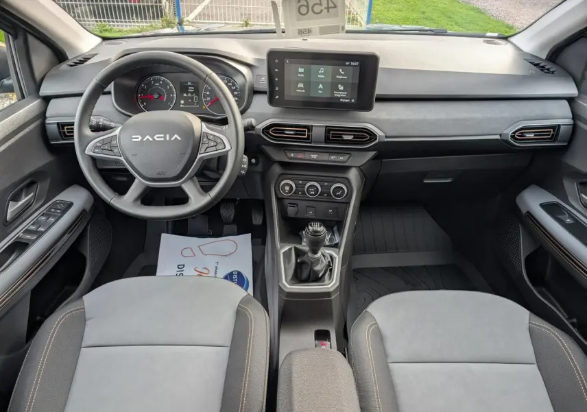 Vue intérieure avant du Dacia Jogger 2025, tableau de bord noir avec volant multifonction et sellerie mixte gris avec surpiqûres brunes.