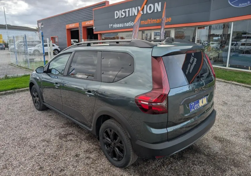 Vue 3/4 arrière droite d'un Dacia Jogger vert métallisé avec jantes noires et barres de toit, stationné devant un garage.
