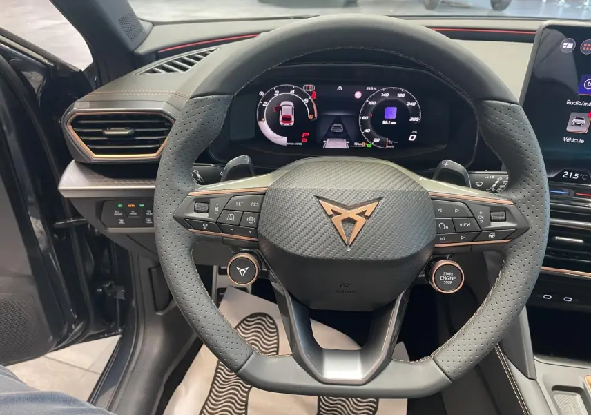 Vue intérieure centrée sur le volant perforé Cupra avec logo cuivré et tableau de bord digital du Leon gris magnetic.