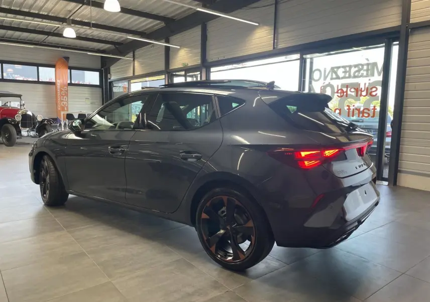 Vue 3/4 arrière droite d'une CUPRA Leon gris magnetic avec jantes cuivrées et feux arrière allumés en intérieur showroom.