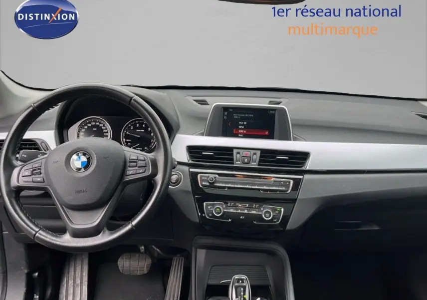 Vue intérieure centrée sur le tableau de bord et le volant noir d'une BMW X1 18i 2018, avec écran multimédia allumé.