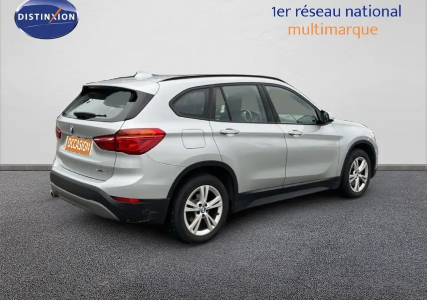 BMW X1 18i gris argent métal vue 3/4 arrière côté droit avec jantes alliage et feux arrière allumés.