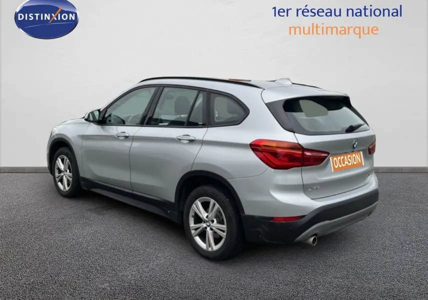 BMW X1 18i gris argent métal en 3/4 arrière droit, avec jantes alliage et feux arrière allumés.
