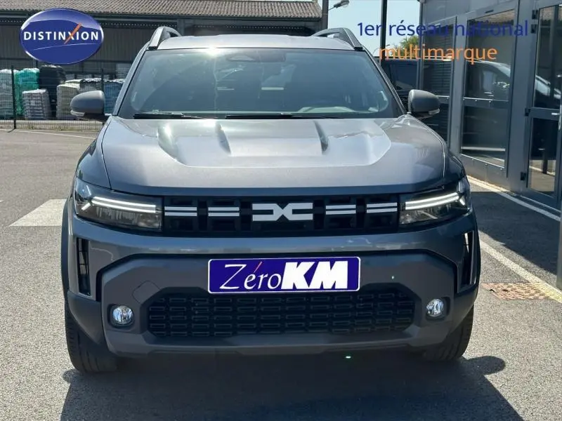 Vue avant d'un Dacia Duster 2025 gris avec calandre noire et phares LED fins, sur parking extérieur.
