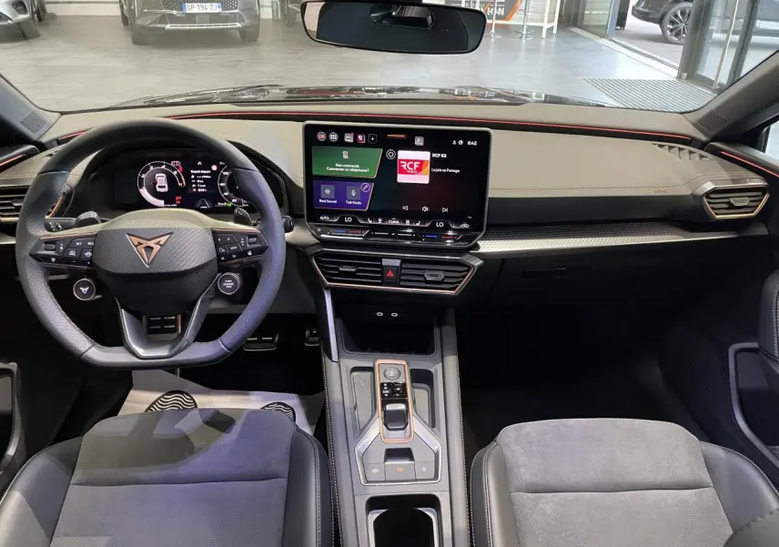 Vue intérieure avant de la CUPRA Leon 2026, tableau de bord noir avec écran tactile central et volant sport à logo cuivré.