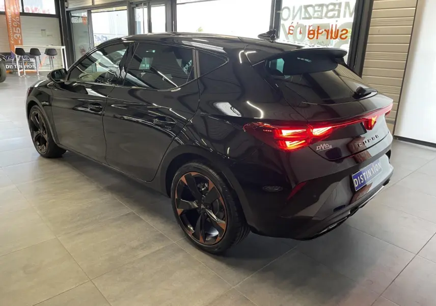 Vue 3/4 arrière droite d'une CUPRA Leon noire avec jantes alliage cuivrées et feux arrière allumés en intérieur showroom.