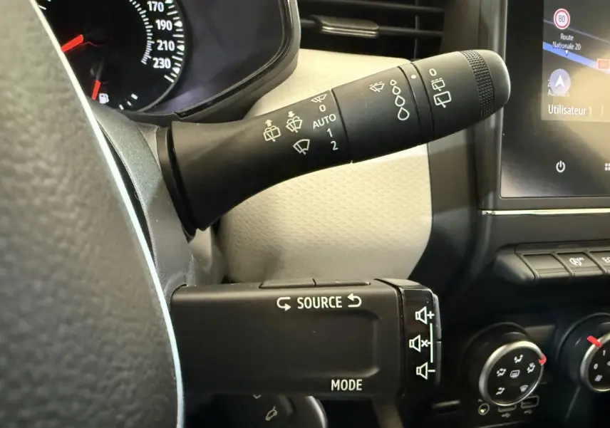 Gros plan sur les commandes au volant et levier d’essuie-glace d’une Renault Clio blanche, tableau de bord partiellement visible.