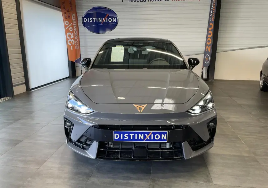 Vue avant d'une CUPRA Leon gris Graphene avec logo cuivré et phares allumés en showroom intérieur.