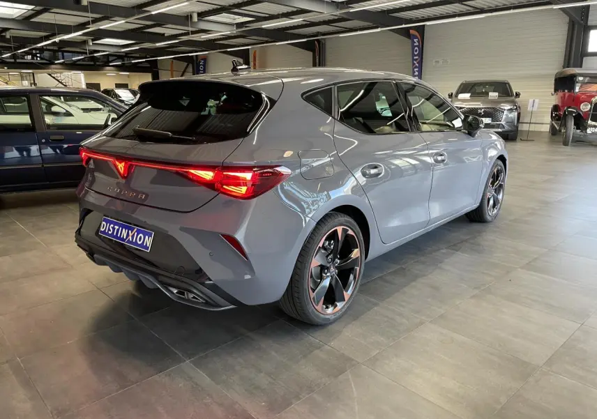 Vue 3/4 arrière droite d'une CUPRA Leon gris Graphène avec jantes cuivrées et feux arrière Tri-LED allumés en intérieur showroom.