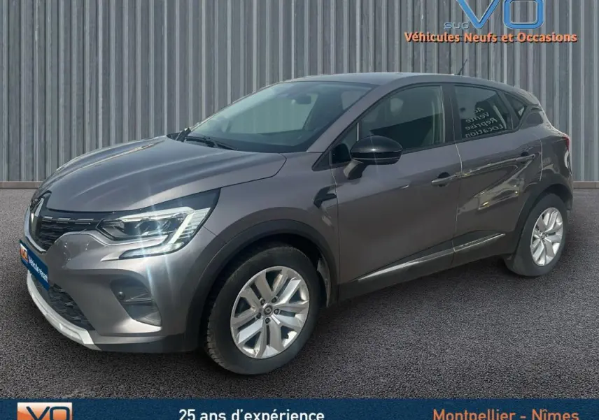 Renault Captur TCe 100 Business gris vu en 3/4 avant droit sur fond industriel, avec feux allumés et jantes argentées.