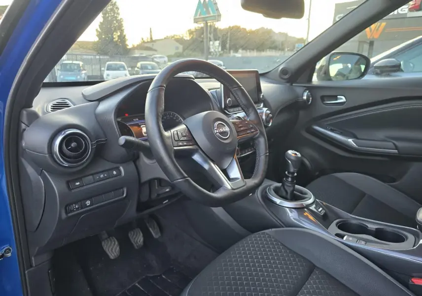 Intérieur avant droit du Nissan Juke II bleu magnetic 2024 avec volant cuir, boîte manuelle et console centrale moderne.