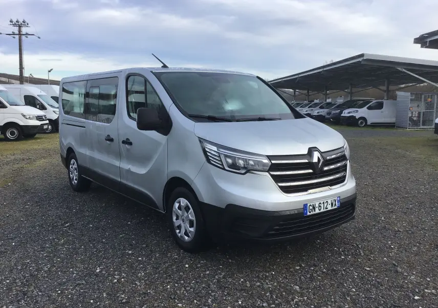 Renault Trafic Combi gris clair métal vu en 3/4 avant droit sur un parking avec plusieurs autres véhicules utilitaires.