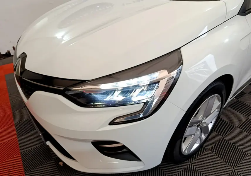 Vue 3/4 avant droit d'une Renault Clio Business blanche avec phares LED allumés et jantes alliage visibles.