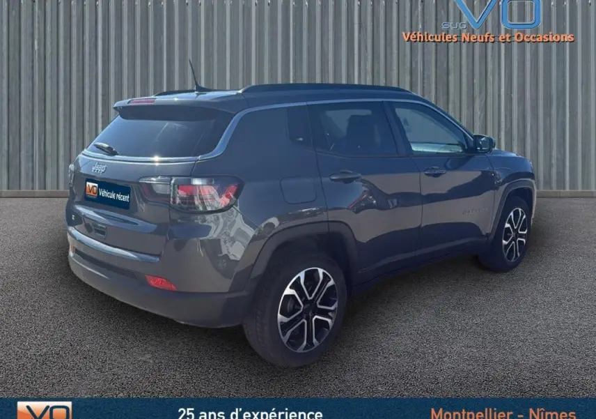 Jeep Compass gris 3/4 arrière droit, avec jantes 18 pouces et barres de toit visibles sur fond urbain.