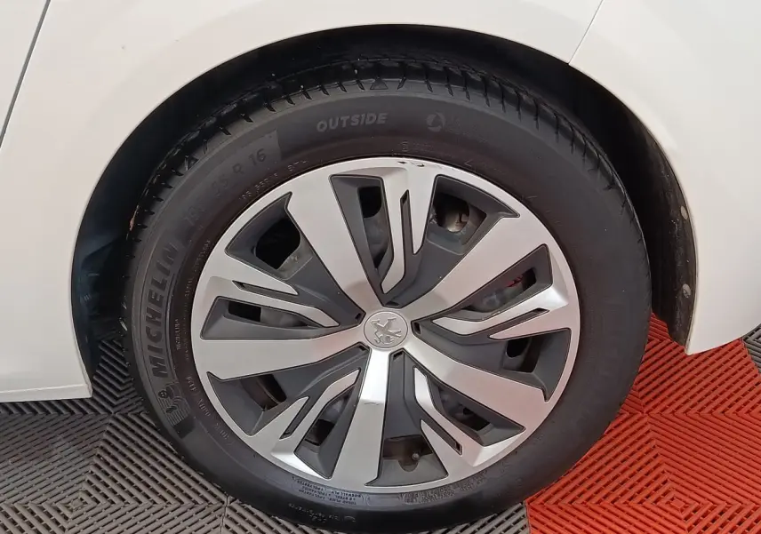 Gros plan sur la roue avant gauche d'une Peugeot 208 blanche, avec jante alliage au design moderne.