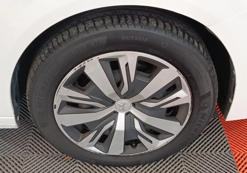 Gros plan sur la roue avant gauche d'une Peugeot 208 blanche, avec jante alliage au design moderne.