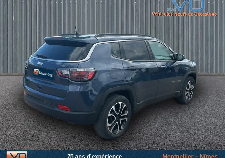 Jeep Compass bleu vue 3/4 arrière droit, avec barres de toit et jantes 18 pouces visibles sur sol goudronné.