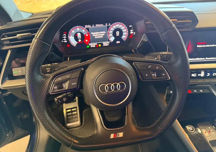 Vue rapprochée du volant cuir perforé noir avec logo Audi et badge S, tableau de bord digital et console centrale visibles.