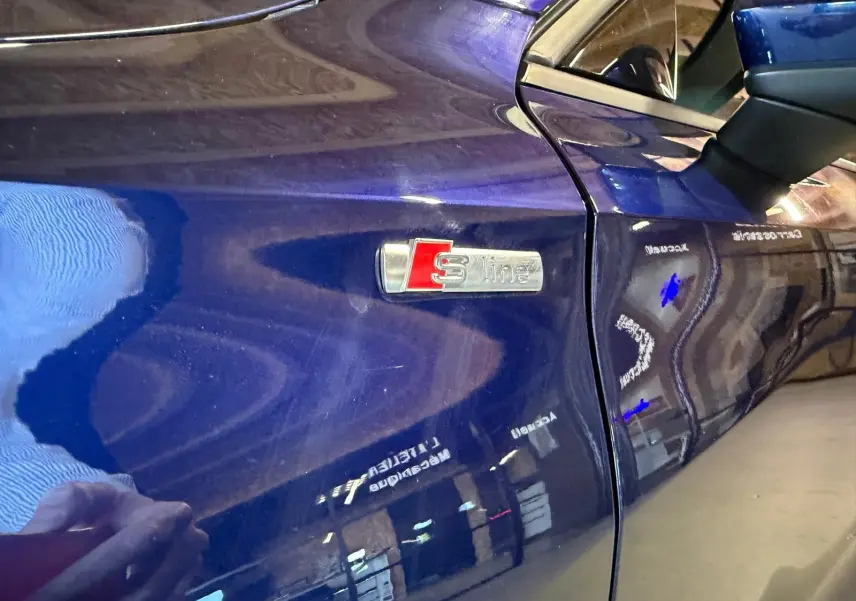 Gros plan sur l'aile avant gauche bleu foncé d'une Audi A3 S-Line, mettant en valeur l'emblème S-Line chromé et rouge.