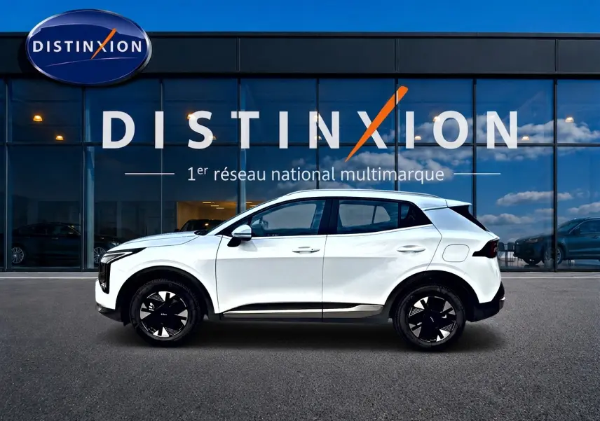 Vue de profil côté gauche d'un KIA SPORTAGE blanc 1.6 hybride 239 active avec jantes alu noires devant une vitrine Distinxion.