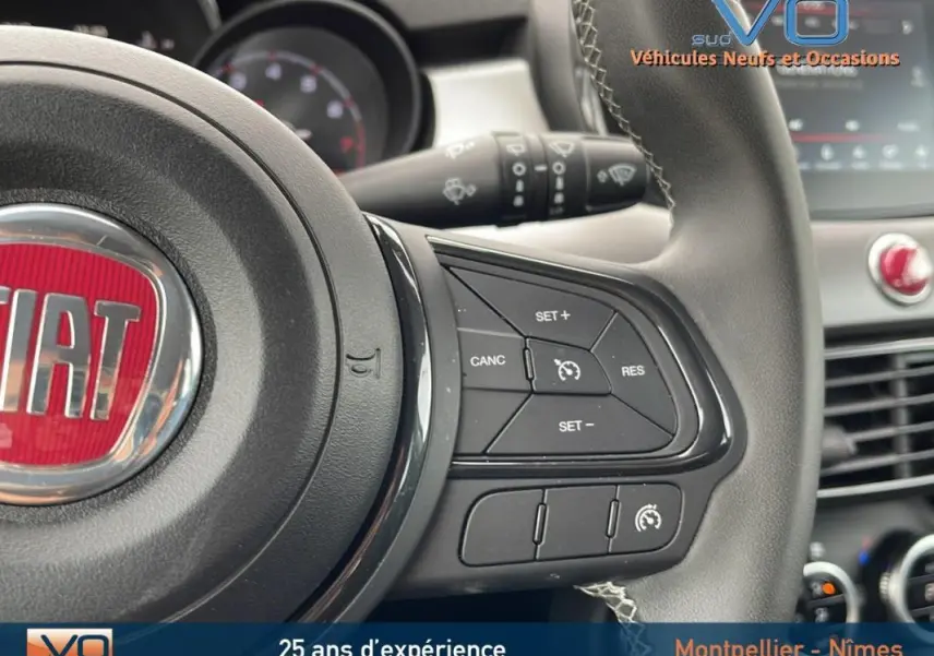 Gros plan sur le volant sport de la Fiat 500X gris, montrant les commandes du régulateur de vitesse et le tableau de bord flou.