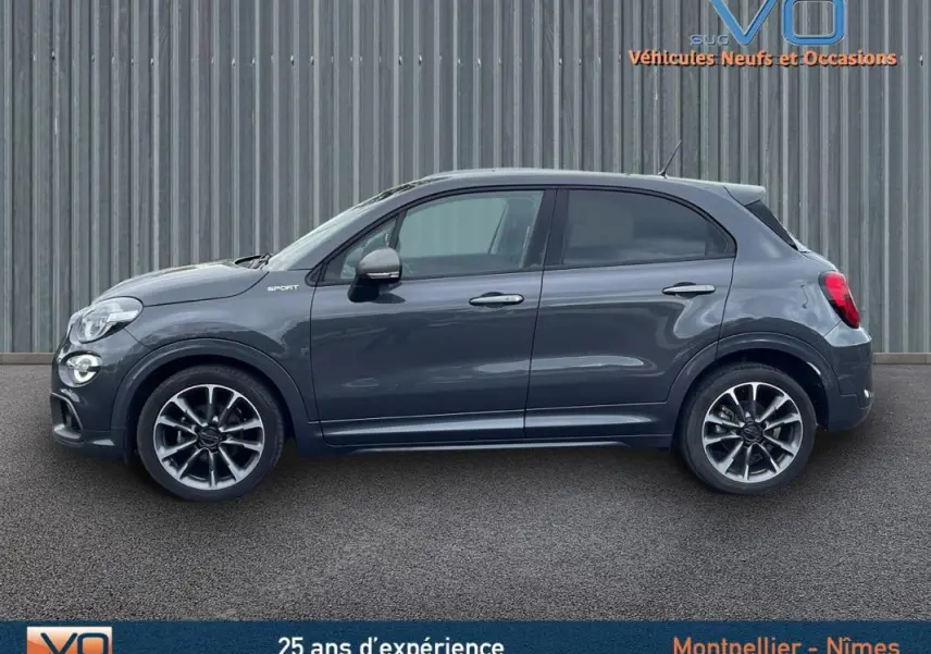 Profil côté gauche d'une FIAT 500X gris métallisé MY22 Sport avec jantes alliage 18 pouces sur fond industriel.