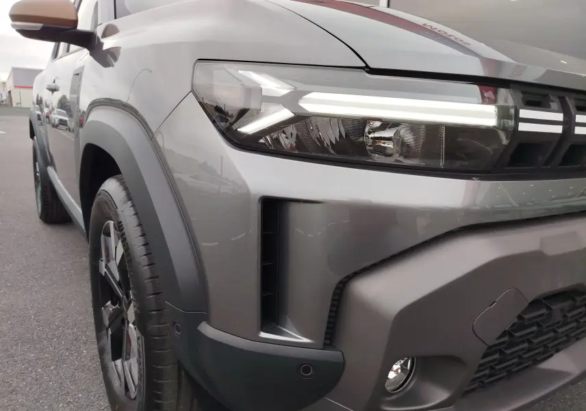 Gros plan sur l'avant droit gris schiste du Dacia Duster 2025, mettant en valeur son phare LED et son bouclier robuste.
