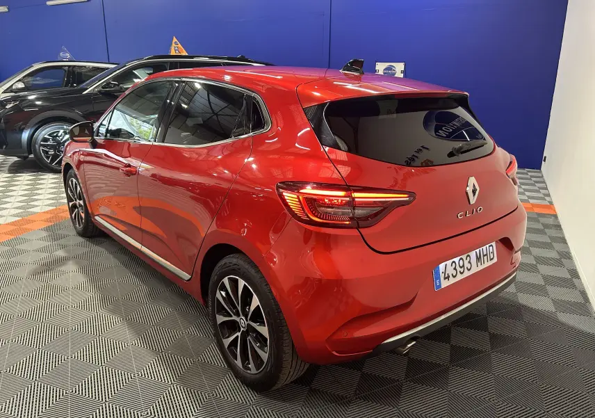 Vue 3/4 arrière droite d'une Renault Clio V rouge 2023 avec feux allumés dans un showroom intérieur.