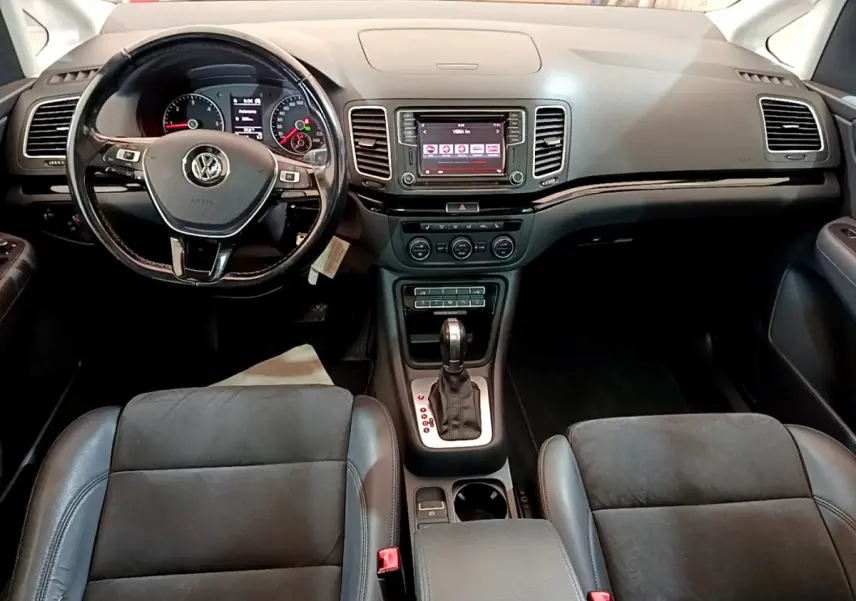 Vue intérieure avant du Volkswagen Sharan 2017 avec tableau de bord noir, volant multifonctions et console centrale avec boîte auto.