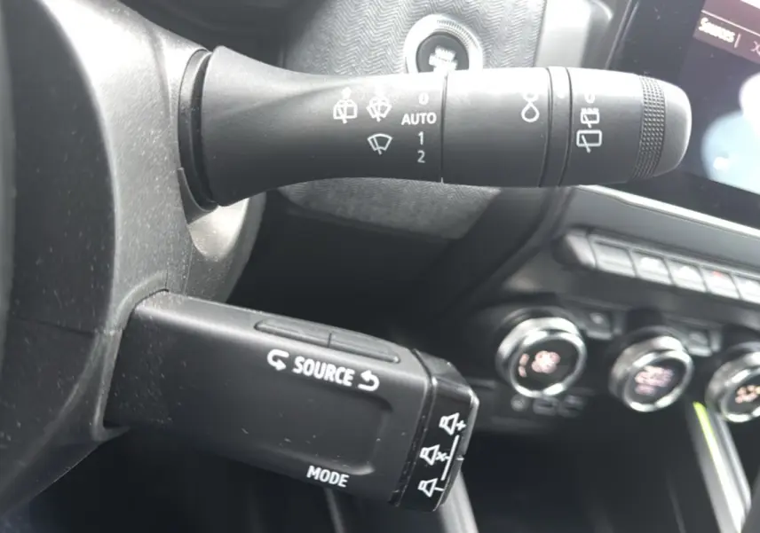 Gros plan sur la commande multifonction gauche du volant de la Renault Clio gris schiste, avec tableau de bord et commandes clim visibles.