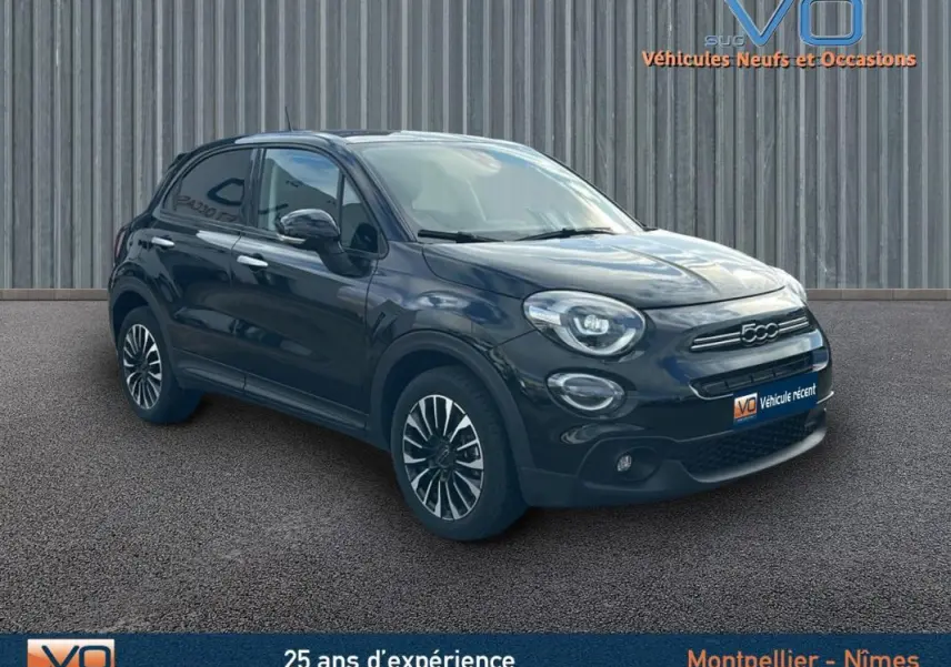 Vue 3/4 avant d'une FIAT 500X noire avec jantes classiques 16 pouces sur un sol goudronné devant un mur métallique.