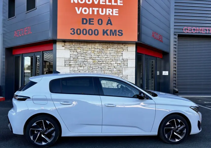 Vue latérale droite d'une Peugeot 308 Hybrid 145 e-DCS6 Allure blanche, garée devant un bâtiment avec panneau orange.