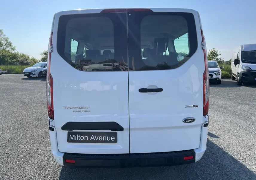Vue arrière d'un Ford Transit Custom blanc avec hayon vitré et logo Ford visible sur un parking.