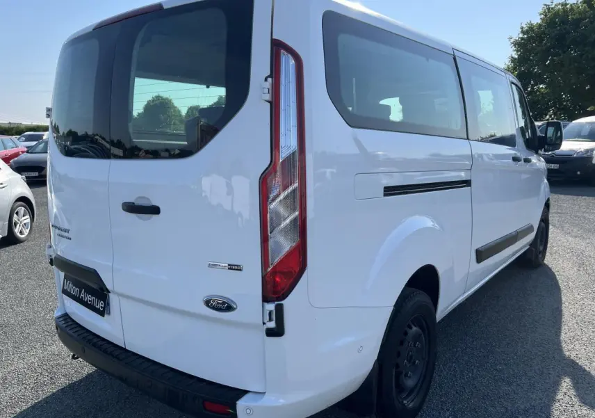 Vue 3/4 arrière droite d'un Ford Transit Custom Kombi blanc avec hayon vitré et badges Hybrid visibles.