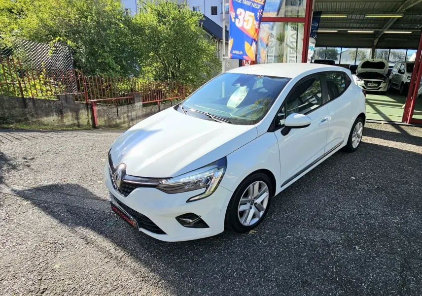 Renault Clio blanc glacier vue 3/4 avant droit, garée devant un garage avec panneaux publicitaires colorés.