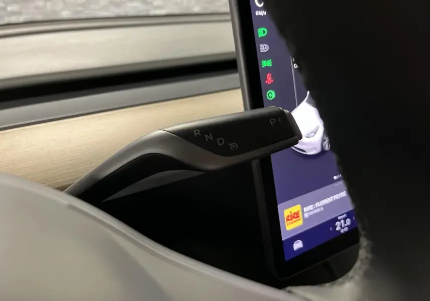 Gros plan sur la commande de boîte automatique noire et argentée, avec l'écran tactile et le tableau de bord bois clair d'une Tesla Model Y blanche.