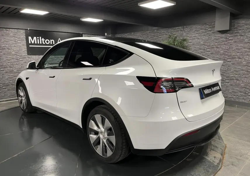 Tesla Model Y blanc nacré vu en 3/4 arrière droit dans un showroom avec toit panoramique noir brillant.