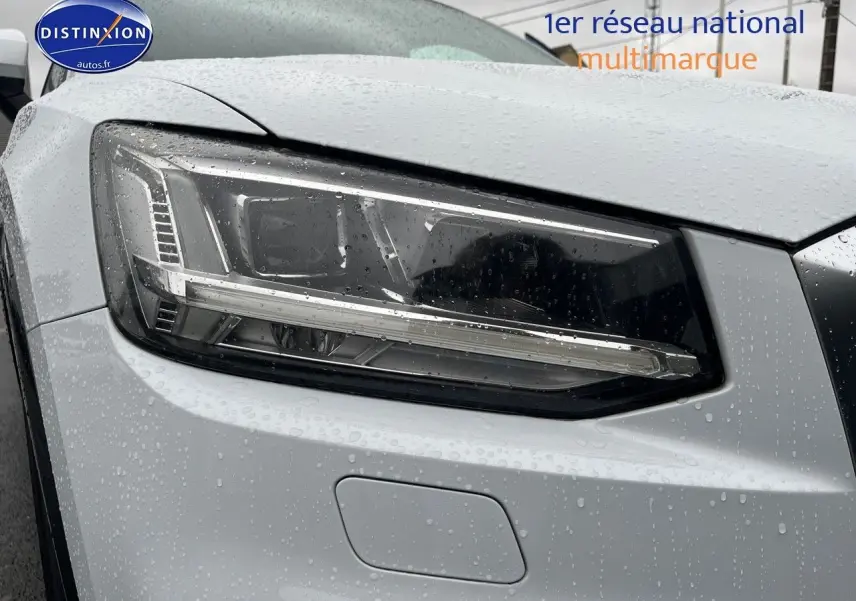 Gros plan sur le phare avant gauche d’un Audi Q2 blanc Ibis avec gouttes de pluie visibles sur la carrosserie.