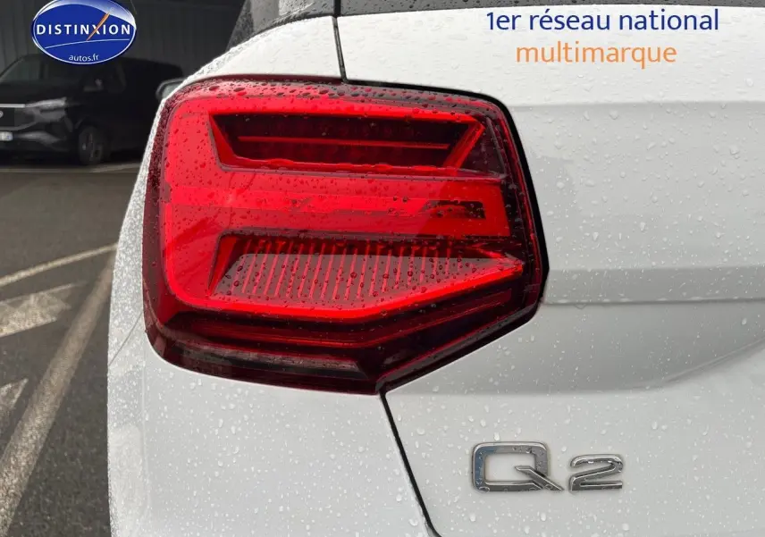 Gros plan sur le feu arrière gauche d’un Audi Q2 blanc Ibis avec logo Q2 visible sous la pluie.
