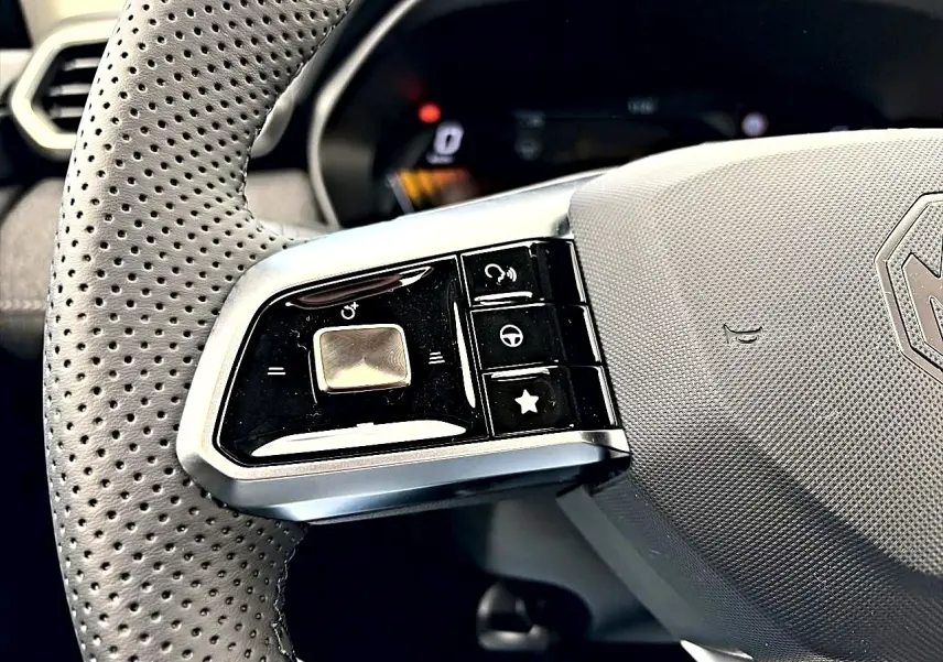 Gros plan sur la partie gauche du volant perforé gris du MG Motor ZS 1.5 hybrid 197 avec commandes intégrées.