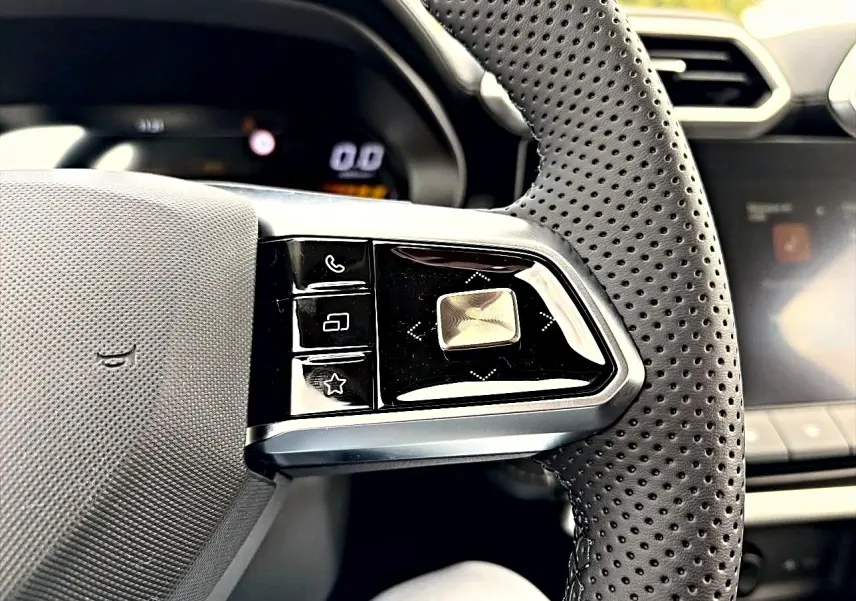 Gros plan sur les commandes gauche du volant perforé noir du MG MOTOR ZS 1.5 hybrid Camden grey, tableau de bord flou en arrière-plan.