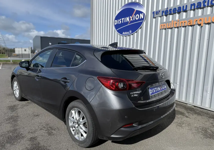 Vue 3/4 arrière droite d'une Mazda3 gris métallisé garée devant un bâtiment avec logo Distinxion visible.