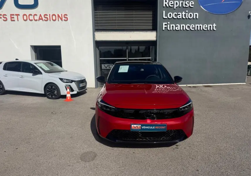 Vue avant d'une Opel Corsa rouge 2025 garée devant un bâtiment avec un panneau publicitaire visible.