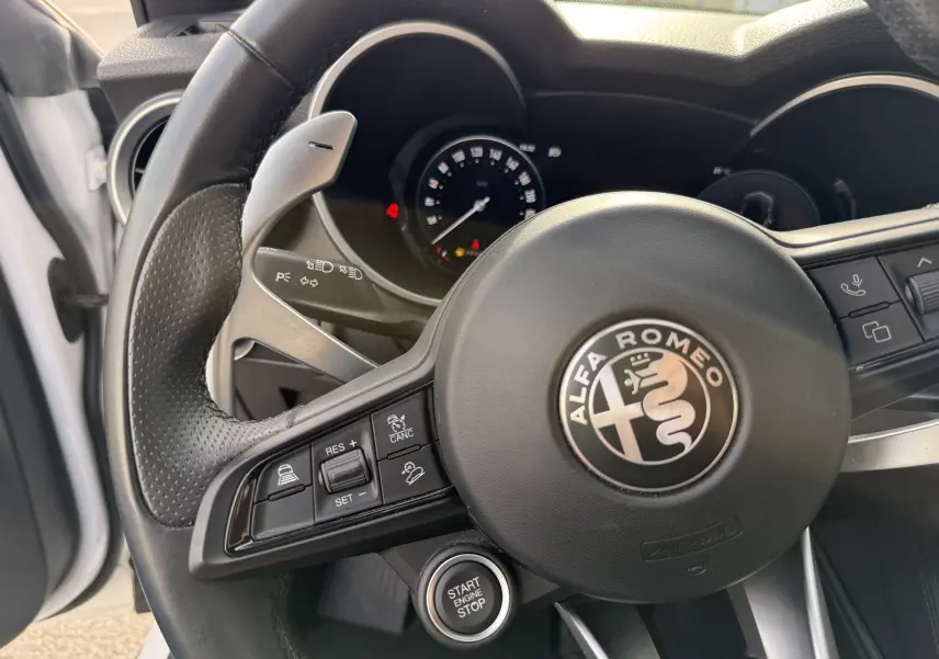 Vue rapprochée du volant noir perforé d'un Alfa Romeo Stelvio blanc, avec commandes intégrées et bouton start/stop visible.